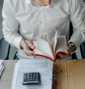 Man in white shirt reading book at office desk with calculator and documents.