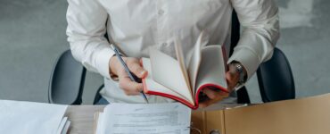 Man in white shirt reading book at office desk with calculator and documents.