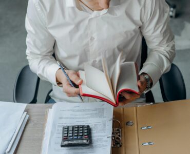 Man in white shirt reading book at office desk with calculator and documents.